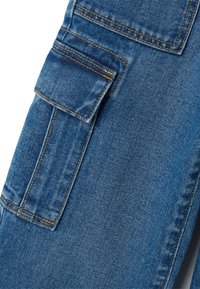 Tela de mezclilla azul con un bolsillo con solapa asegurado por costuras, con hilo de color naranja-marrón en una sección de la prenda.