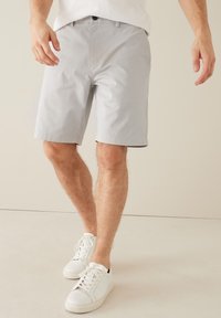 Hellgraue Shorts mit glatter Textur, ausgestattet mit einem Knopfverschluss und Taschen, kombiniert mit weißen Turnschuhen auf einem neutralen Hintergrund.