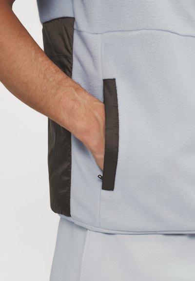 Lyseblå fleecejakke med sorte nylon detaljer, der har en sidelomme. Teksturen virker blød med en kontrasterende glat overflade.