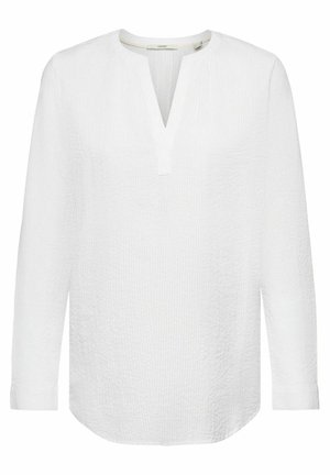 Persona con gabardina beige, camiseta blanca, pantalones anchos blancos y sandalias marrones sobre un fondo azul.