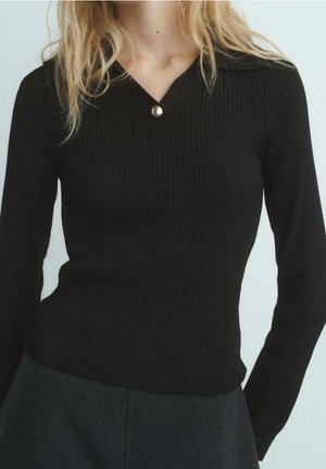 Femme portant un pull noir ajusté en côtes, manches longues, avec un petit bouton rond doré au col en V et un pantalon gris foncé.