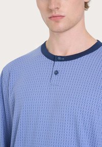 Camicia azzurra a maniche corte con motivo di ancore, colletto girocollo e bordi blu scuro. Presenta un'apertura con patta a due bottoni e una texture di tessuto morbido.