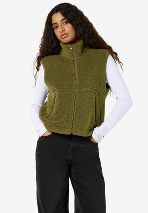 Jonge vrouw met lang krullend haar die een groene fleecevest draagt over een witte lange mouwen top en een zwarte broek, staand met handen in de zakken.