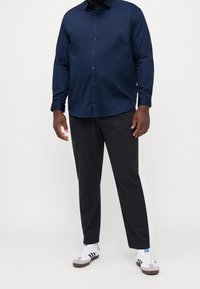 Camicia blu navy con bottoni e maniche lunghe, abbinata a pantaloni scuri e sneakers bianche con strisce nere e accenti grigi.