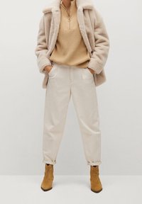 Beige faux fur frakke over en tan striktop og cremede bukser. Brune ruskinds ankelstøvler fuldender looket. Bløde teksturer og neutrale farver.