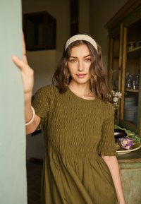 Ženska z valovitimi rjavimi lasmi in bež trakcem za glavo, oblečena v olivno zeleno obleko, stoji v zaprtih prostorih blizu lesene omare s krompirjem na krožniku.