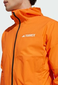Veste imperméable orange avec fermeture éclair à l'avant, capuche élastique et accents noirs. Elle présente le logo "TERREX" en blanc sur la poitrine.