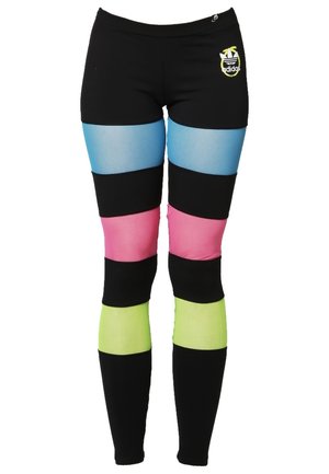 Žena stojí s rukou na boku, má na sobě bílý krátký top s nápisem "PROČ?" a černé legíny s modrými, růžovými a žlutými barevnými bloky.