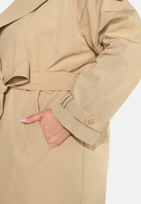 Beżowy trencz związywany w talii, z kieszeniami i detalami przy mankietach, o gładkiej teksturze i minimalistycznym wzornictwie.