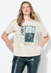 Beżowa koszulka bawełniana z zielonym grafiką słońca nad falami i wzorami kwiatowymi, z napisem "Spotkajmy się pod słońcem". Luźny krój.