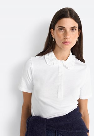 White short-sleeve polo shirt featuring a ruffled collar, button front, and soft texture. A navy sweater is wrapped around the model's waist.
