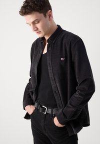 Camisa de pana negra con botones frontales y pequeño logotipo bordado, llevada sobre una camiseta sin mangas negra y combinada con pantalones negros y un cinturón de cuero.