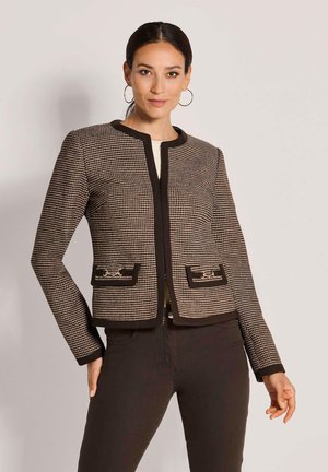 Vrouw met donker haar in een bruin-zwarte jas met patroon en gouden kettingdetail op de zakken, zwarte broek dragend, staand tegen een effen muur.