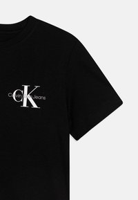 Svart bomulls T-shirt med en vit CK-logotyp och texten "Calvin Klein Jeans" på vänster sida, korta ärmar och rund hals.