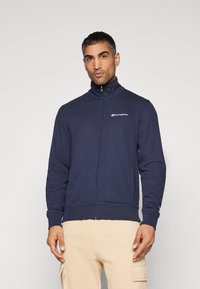 Námornícky zip-up mikina vyrobená z jemného materiálu, s vysokým golierom a vyšitým logom na hrudi. Priliehavé manžety a pás.