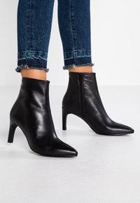 Stivali neri in pelle fino alla caviglia con punte a punta e tacchi sottili indossati con jeans blu a orlo sfrangiato e cropped su un pavimento bianco.
