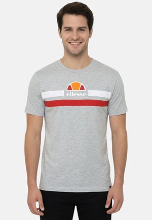 Homme souriant portant un T-shirt gris clair avec des rayures blanches et rouges et un logo en demi-cercle orange avec le texte « ellesse », debout devant un fond blanc.