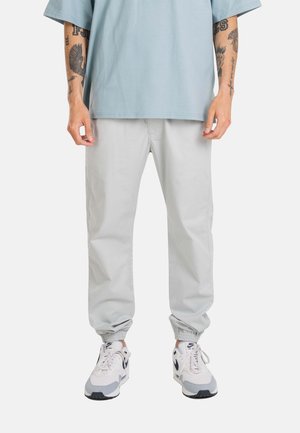 Person wearing light grey jogger pants, white Nike Air Max sneakers, and a light blue oversized short-sleeve shirt, with tattooed arms visible.