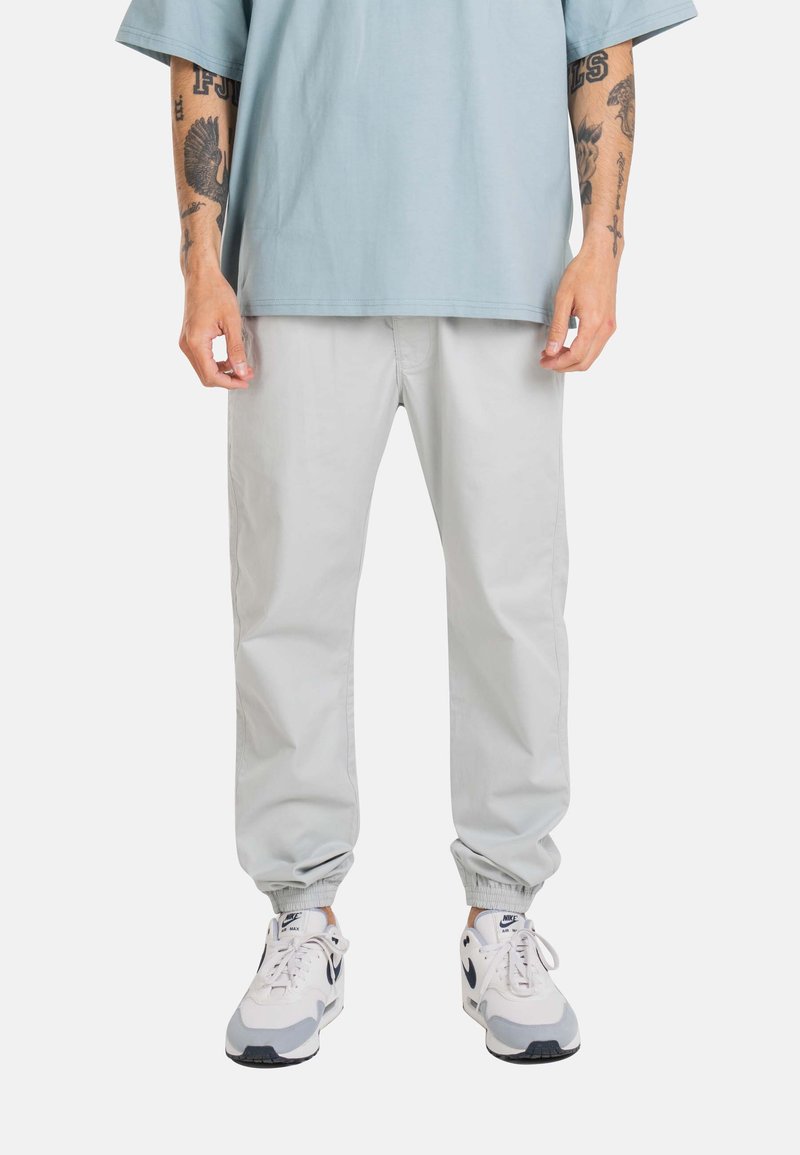 Persona que lleva pantalones jogger gris claro, zapatillas blancas Nike Air Max y una camisa de manga corta azul claro y de gran tamaño, con los brazos tatuados visibles.