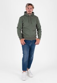 Felpa verde con cappuccio e lacci, abbinata a jeans in denim blu e sneakers bianche. Design semplice, texture morbida e vestibilità rilassata.
