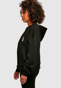 Sudadera con cremallera negra, hecha de un tejido suave, presenta un corte holgado, capucha y puños acanalados. Un pequeño gráfico está bordado en el pecho.