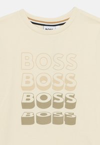 Κρεμ μπλουζάκι με στρογγυλή λαιμόκοψη που φέρει στοιβαγμένο κείμενο "BOSS" σε διάφορες αποχρώσεις του μπεζ και του γκρι, τονίζοντας ένα έντονο, γραφικό σχέδιο.