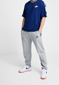 Námořnická modrá tričko s bílým logem, šedé tepláky s černým brandingem a bílé tenisky s červenými detaily. Ležérní sportovní outfit.