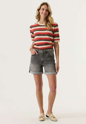 Smiling woman with long blonde hair wearing a red, brown, and beige striped short-sleeve top, rolled black denim shorts, and beige loafers.