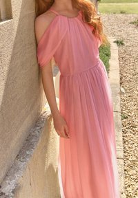 Ljusrosa golvlång klänning med en flödande textur, off-shoulder-design och samlad midja, placerad mot en stenmur och grusväg.