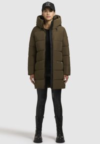 Lang, pufferjakke i olivengrøn med hætte, udstyret med lynlåslofter og en frontlynlås, parret med sorte leggings og kampstøvler.