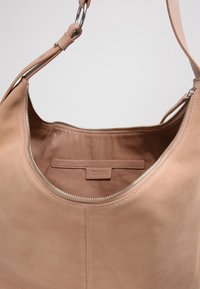 Béžová kožená kabelka s hladkou texturou, která má široké otevírání, stříbrný zip a jemně zaměrou podobný povrch na vnější straně.