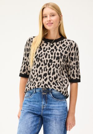Lyshåret kvinde iført en beige og sort leopardmønstret kortærmet sweater og blå cowboybukser, stående mod en lys baggrund.