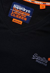 Camiseta negra de cuello en V con el logo bordado "SuperDry" en gris claro. Presenta una etiqueta naranja que dice "Superdry The Orange Label Tee" y tamaño "Mediano."