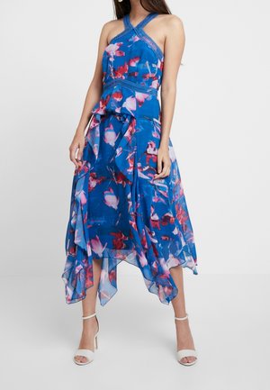 Blaues Blumenkleid mit Neckholder-Ausschnitt, mehrlagigem Design, transparentem Stoff und pinken Akzenten. Kombiniert mit weißen Sandalen mit Absatz.