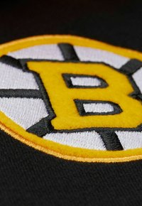 Schwarzstoff-Patch mit einem gestickten kreisförmigen Design und einem weißen Rand, gelbem "B" in der Mitte, umgeben von abwechselnden schwarzen und weißen Streifen.