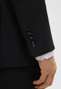 Manica di giacca nera con quattro bottoni neri e dettagli di cucitura. Polsino di camicia bianca visibile sotto l'apertura della manica.