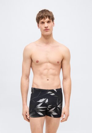 Fiatal férfi áll, ing nélkül, fekete fürdőnadrágban, nagy szürke levélmintákkal, egyszerű, világos háttér előtt.