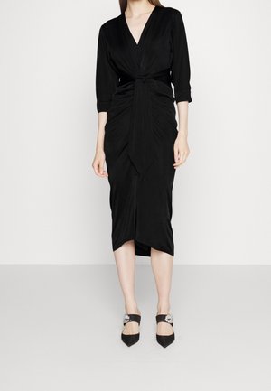 Žena v černých šatech po kolena s tříčtvrtečními rukávy, s vpředu zavazovacím detailem, a černých špičatých botách s ozdobnými stříbrnými přezkami.