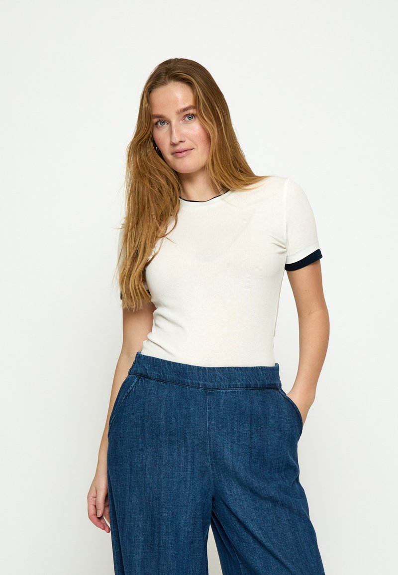 Žena s dlhými svetlohnedými vlasmi, oblečená v bielom tričku s krátkym rukávom a čiernym lemovaním, a voľných modrých rifľových nohaviciach, s rukami v kapse.