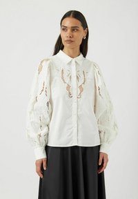 Hvit bluse med lange, puffede ermer som har broderte detaljer i blonder og en klassisk krage. Knapp foran og teksturerte detaljer.
