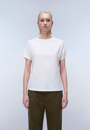 Frau mit kurzem blondem Haar steht frontal und trägt ein schlichtes weißes T-Shirt sowie olivgrüne Hosen vor hellgrauem Hintergrund.