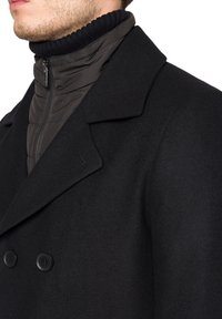 Czarny płaszcz z wełny z podwójnym zapięciem, płaskim kołnierzem i widocznymi czarnymi guzikami; ma ciemną pikowaną wewnętrzną warstwę oraz zamek błyskawiczny.