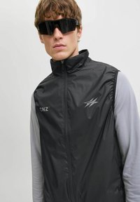 Zwarte lichtgewicht gilet met hoge kraag, ritssluiting aan de voorkant en een grijze langemouwen shirt. Heeft het "CNZ" logo en een strakke, gladde textuur. Zwarte zonnebril gedragen.