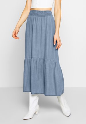 Kobieta ubrana w niebieską, warstwową spódnicę maxi z wysokim stanem i marszczonym paskiem oraz białe, teksturowane botki do kostek.