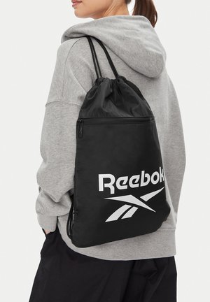 Persona che indossa una felpa con cappuccio grigia e uno zaino con coulisse nero Reebok con logo bianco sul retro, rivolta di spalle rispetto alla fotocamera.