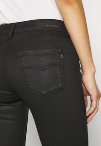 Vaqueros de denim negro con un diseño ajustado. Presenta un bolsillo trasero texturizado con un patrón de costura en forma de chevron y un parche de cuero con marca en la cintura.