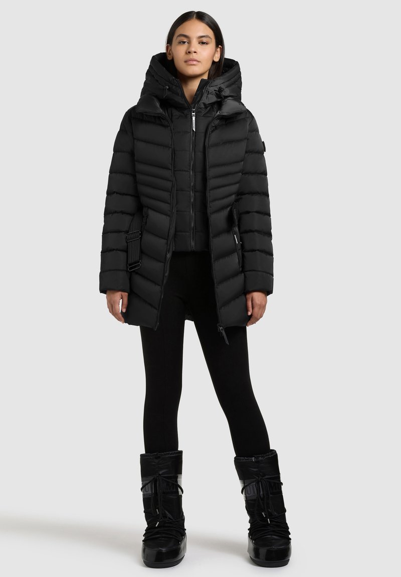Sort dunjakke med høj krave, lodret syning og justerbar hætte. Parret med sorte leggings og vandtætte støvler.