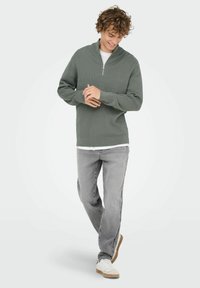 Suéter verde con cuello de cremallera y textura acanalada, combinado con jeans gris claro y zapatillas blancas. El conjunto presenta un look en capas con una camisa blanca debajo.