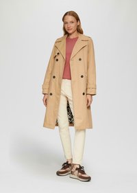 Beige Trenchcoat mit schwarzen Knöpfen, ausgestattet mit einem Futter im Leopardenmuster. Kombiniert mit einem pinken Oberteil, hellen Jeans und braunen Sneakers.