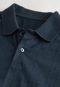 Mørkeblå polo skjorte med et tekstureret ternet mønster, en mesh-krave og en to-knap strop med detalje af en enkelt knap.